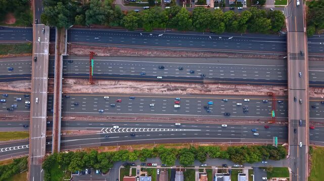 Rising over four multi-lane highways with two overpasses above. Many cars go by the roads at daytime. Residential neighborhoods and woods around the motorways.