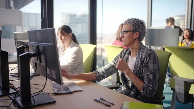 Middle aged professional works at an open plan desk in a bright corporate office, concentrating on tasks at a dual monitor setup with paperwork and notes by the window overlooking the city.