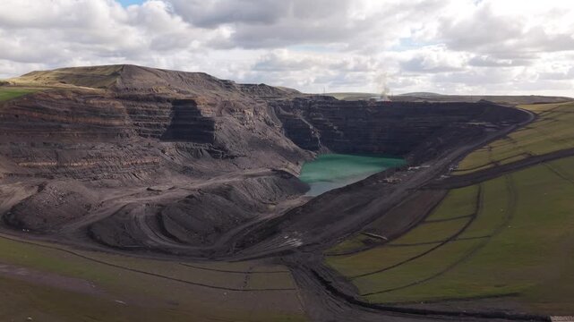 Wide cinematic view across a vast open pit quarry in Wales, revealing deep terraced rock walls, winding access roads and a turquoise lake at the bottom, surrounded by green countryside under a cloudy 