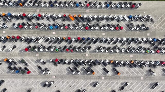 Aerial view of rows of parked new trucks and trailers in a large paved parking lot with surrounding trees and roadway. Drone footage of warehouse of manufactured lorry cars. Industrial concept