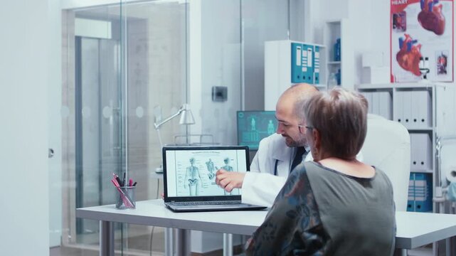 Doctor and older woman study a human skeleton diagram on a laptop during a medical consultation, illustrating anatomy education and discussion of bone health, supporting patient care.