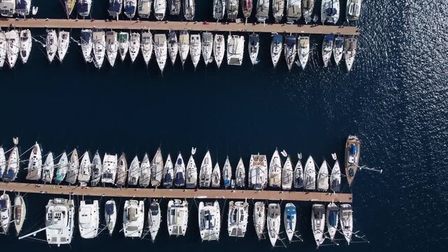 Aerial symmetric pier with aligned yachts peacefully positioned near cityscape features. Calm dark water and luxury sailboats moored at a Turkish dock filmed at clear summer day. Water transport theme