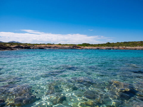 Une vue panoramique sur une baie aux eaux turquoise peu profondes &agrave; Majorque, encadr&eacute;e par une c&ocirc;te rocheuse sauvage et un ciel bleu clair, id&eacute;ale pour illustrer un paradis estival m&eacute;diterran&eacute;en ensol