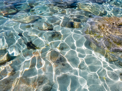 Un gros plan captivant de l'eau cristalline d'une crique majorquine, montrant les reflets dansants du soleil sur les galets immerg&eacute;s, &eacute;voquant une puret&eacute; absolue et la fra&icirc;cheur apaisante d'une matin&eacute;