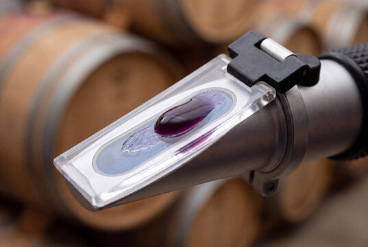 A drop of red grape must on the glass prism of a handheld refractometer inside a wine cellar with wooden barrels.