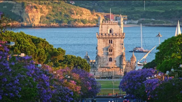Blooming jacaranda trees frame Belem Tower in Belem, Lisbon, Portugal. Tourist boats float on the water as the sun sets, creating a beautiful view of the area filled with purple flowers.