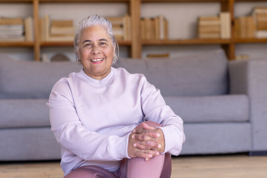 Senior African woman sitting on hardwood floor by grey sofa in living room in pastel top
