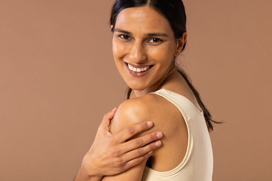 Woman turning toward camera, smiling and holding shoulder in studio wearing beige tank top
