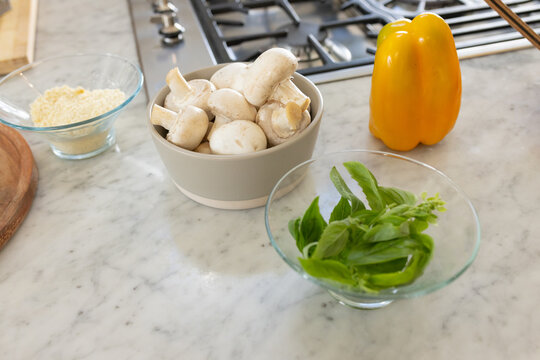 Neutral ceramic bowl is holding white mushrooms on marble countertop near basil yellow pepper tongs