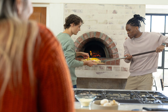 Diverse friends handling pizza from wood-fired oven in kitchen, mauve top man using long peel