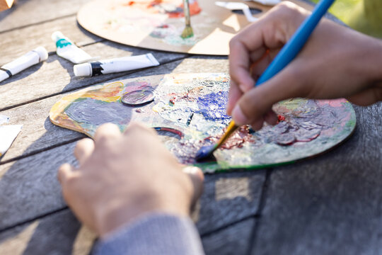 Man mixing blue paint with blue brush on palette at wooden table, sweater cuff showing, tubes