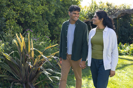 Diverse couple walking hand in hand through lush garden, passing pointed leaf plant and pergola
