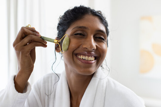 Smiling Indian woman rolling jade facial roller on right cheek in bathroom wearing white bathrobe