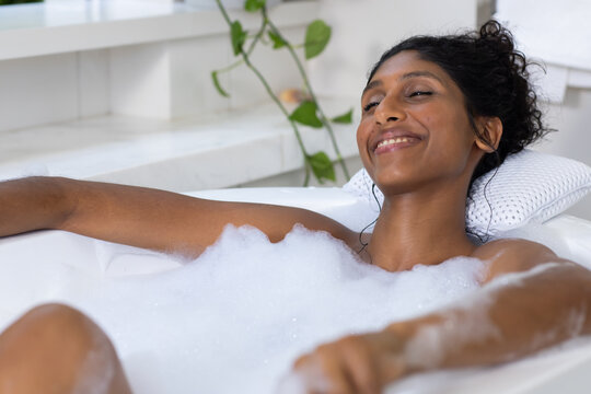 African American woman reclining in bathtub soaking in foam, resting on pillow, wearing earphones