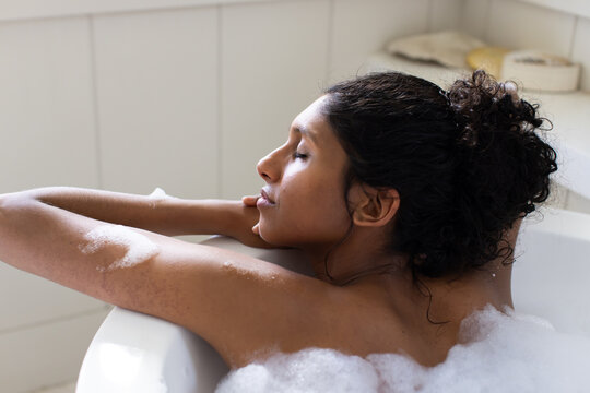 Woman reclining in foamy bathtub in bathroom, light coming from left, towels and soap on ledge