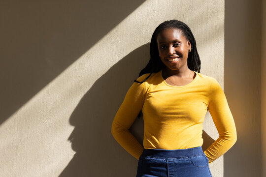 African American woman leaning on wall angled sun casting shadow, mustard top dark pants