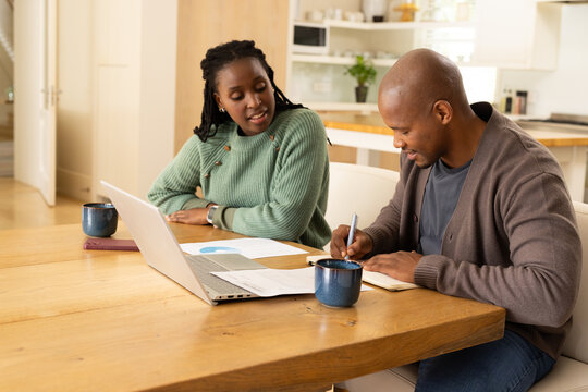 African couple sitting at wooden dining table reviewing laptop, woman in green sweater, man writing