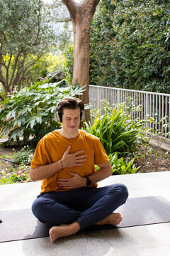 Sitting crosslegged 20s man meditating on patio yoga mat wearing orange tee headset wristwatch