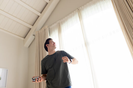 Man in 20s standing at home by sheer curtains holding phone notebook listening with wired earphones