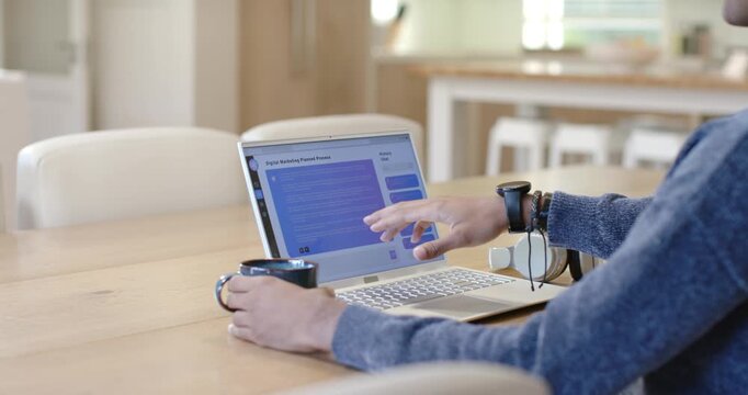 Noticing blue form, woman is tapping rows on laptop to review, holding coffee at kitchen table