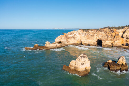 Coastal rock formations and a sea cave line the shoreline, with blue ocean water and clear sky.