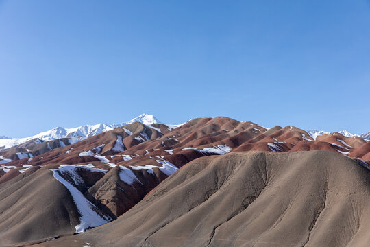 Majestic red clay and brown sedimentary mountain ridges with snow patches in Osh Region under bright clear blue sky, showcasing the rugged natural wild landscape of Kyrgyzstan
