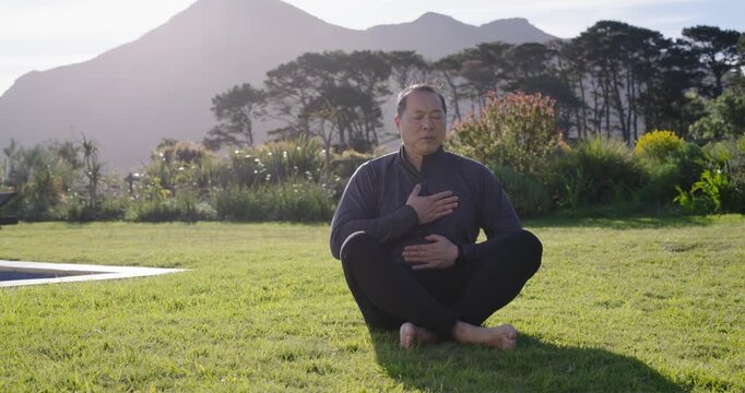 Asian man crosslegged beside pool holding hands on chest and belly, starting breathing to calm mind