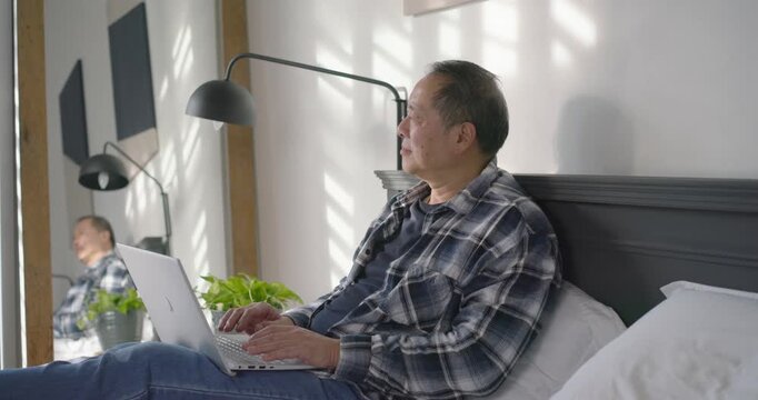 Mature Asian man settling on bed, typing on laptop, glancing at mirror and working