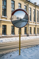 Fototapeta premium Round traffic mirror reflects snowy city street, parked car. Yellow pole stands on sidewalk near yellow brick building. Winter road with snow, ice covers ground. Urban landscape shows cold weather