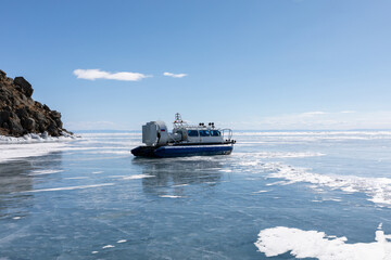 Transport hovercraft vehicle moving on thick blue ice of frozen Lake Baikal in Irkutsk Oblast, Russia under sunny winter sky with rocky cliff shore visible in background distance © TravelFlow