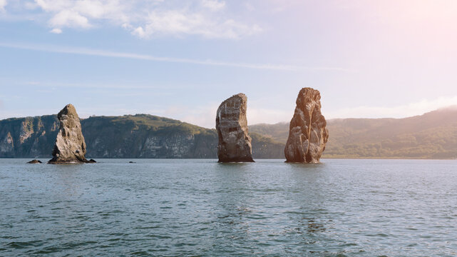 Scenic seascape of the famous Three Brothers basalt sea stacks rising from Avacha Bay waters against green coastal cliffs in Kamchatka Krai, Russia during a sunny summer day