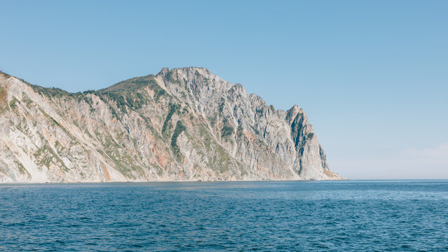 Rocky coastal mountain ridge rising steeply from the calm sea under a clear blue sky, capturing the wild untouched beauty of nature in Kamchatka Krai, Russia on a sunny day