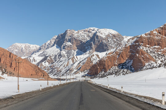 Empty asphalt road stretches toward snow covered peaks and red rock formations under a clear blue sky across the scenic winter landscape of Osh region in Kyrgyzstan