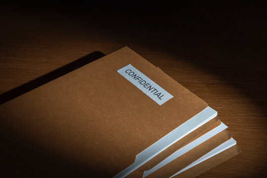 Stack of brown manila folders labeled CONFIDENTIAL on a wooden desk. Corporate secrecy, classified documents, legal privacy concept.