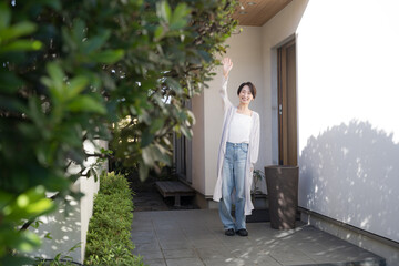 おしゃれで綺麗な新築一戸建ての緑あふれる玄関先で、笑顔で見送る日本人の妻、いってらっしゃいと挨拶する朝の風景・全身イメージ © kapinon