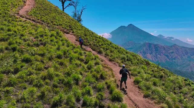 Aerial view of two trail runners ascending a steep mountain ridge path with Mount Rinjani backdrop