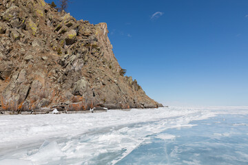 Scenic view of a rugged rocky cliff rising above the vast frozen surface of Lake Baikal, covered with ice hummocks and clear blue ice beneath a bright clear blue winter sky in Siberia © TravelFlow