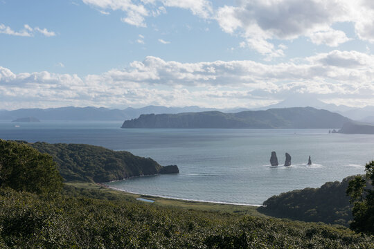 Panoramic high-angle view over Avacha Bay with Tri Brata sea stacks, green headlands, mountain silhouettes, and blue waters in Kamchatka Krai, Russia under scattered clouds