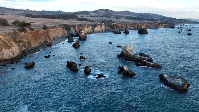 Drone view of towering sea stacks and coastal cliffs near Duncan's Point in Bodega Bay, California, featuring crashing ocean waves, waterfront homes, and scenic rolling hills.