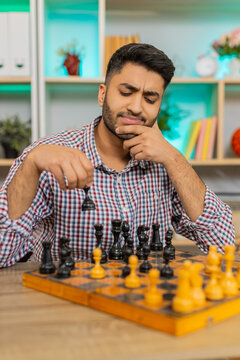 Indian man on home table plays chess and studies next move with calm smile and deep focus during board game. Arabian guy on home table thinks over strategy and enjoys calm leisure with brain training.