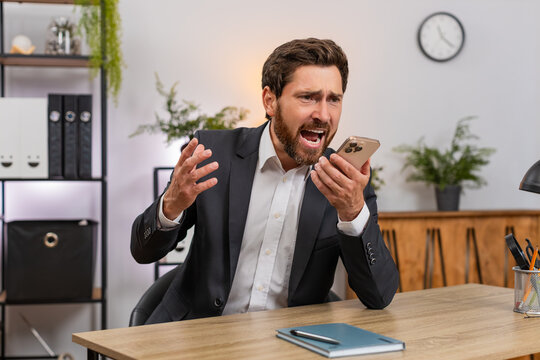 Middle-aged businessman at home office argues on smartphone speaker shouting very waving hand during call. Freelancer guy at table ends conversation sighs angrily staring at device feeling frustrated.