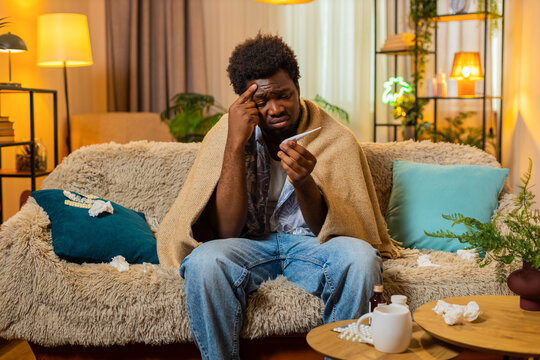 African American young man on home sofa checks thermometer with worried face under blanket during illness. Black guy on home sofa feels sad about fever and chills while resting sick indoors at home.