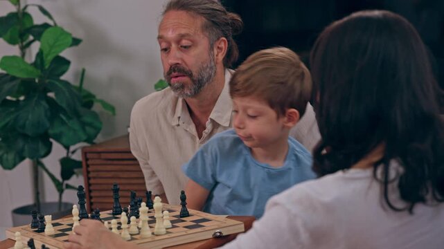Father and boy play chess board game at home as family shares quiet moment of learning, concentration, and connection around table with child and another person watching