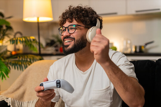 Arabian man at home plays on game console holds joystick leans forward enjoys play sees level score high. Egyptian guy on sofa cheers after win presses buttons shoots jumps feels thrill night victory.