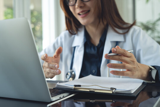 Smiling female doctor talking during online consultation while reviewing medical documents at laptop in clinic office, telemedicine concept