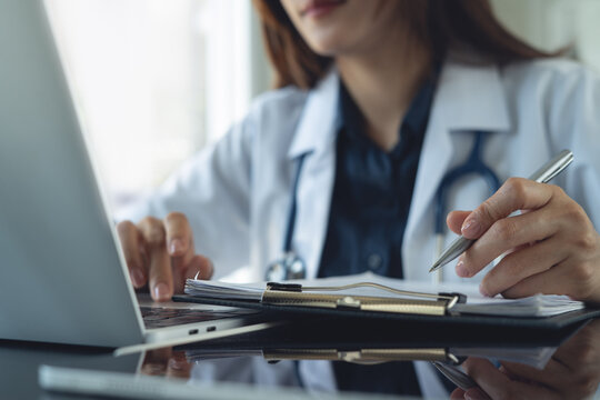 Close up of female doctor working on laptop and writing in medical documents on clipboard at desk n clinic office.