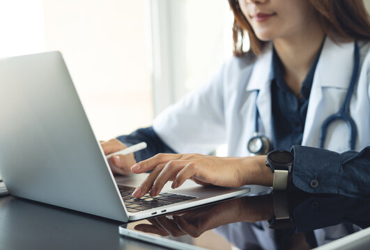 Doctor typing on laptop, managing patient data with digital tablet on desk in clinic office.