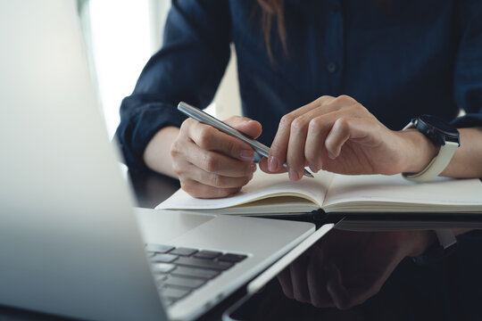 Close-up of hands holding a pen over an open notebook beside a laptop on office desk, concept of planning, note taking, productivity and business work.
