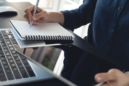 Close-up of hand writing note in notebook beside laptop at desk, planning ideas, working in modern office