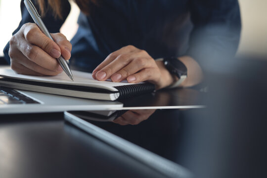 Close-up of woman writing notes in a notebook beside laptop and tablet at desk, planning work and organizing ideas in a modern workspace.